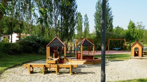 A playground with wooden play structures, including a swing set and a climbing area, surrounded by sand and greenery.