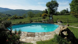 A swimming pool with a stone deck, surrounded by a wooden fence, set in a lush green landscape with hills in the background.