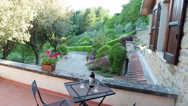 A terrace with a table and chairs, a bottle of wine, and a view of a garden with trees and a pathway. - Villa in Casentino with Pool & Castle Views (Stia)