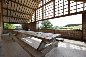 Un pavillon en plein air doté d’une table et de bancs en bois, de grandes fenêtres et offrant une vue sur la campagne.