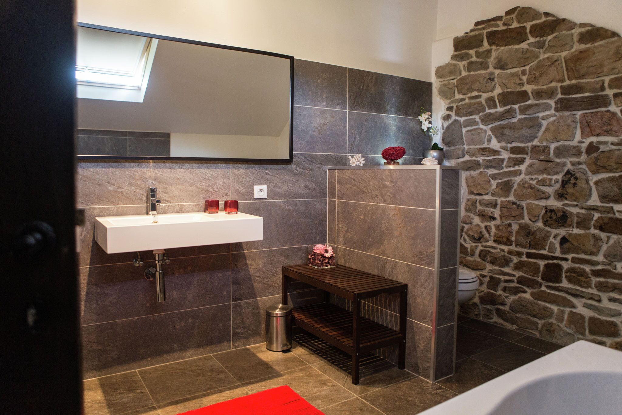 A modern bathroom with a stone wall, a white sink, and a small wooden shelf with decorative items.