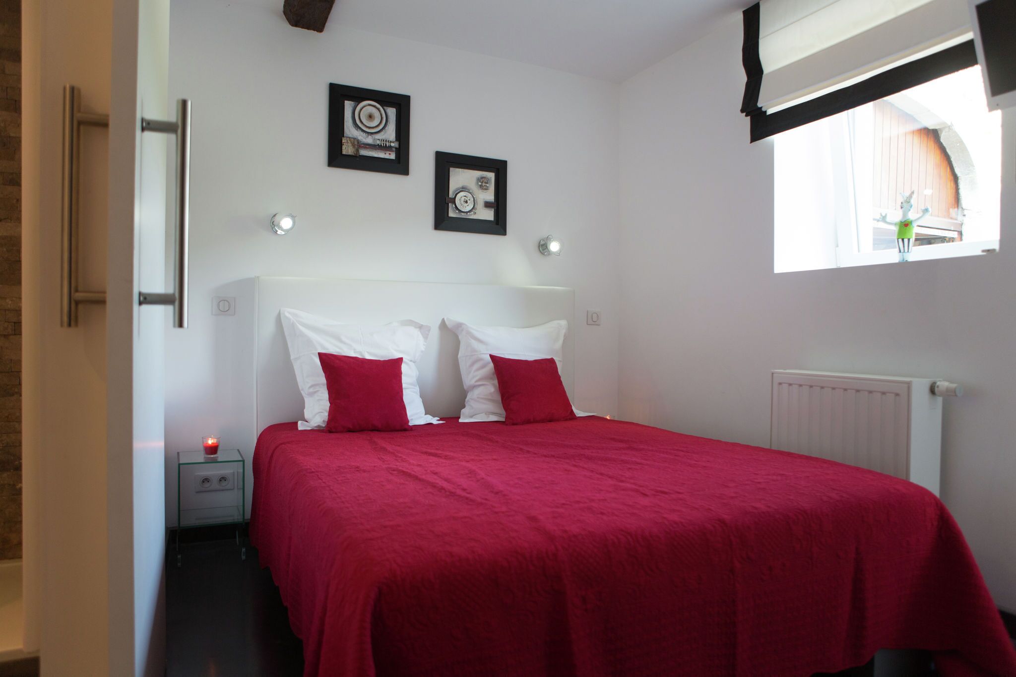Una camera da letto con un letto, due cuscini rossi, un comodino con una candela e due quadri appesi al muro.