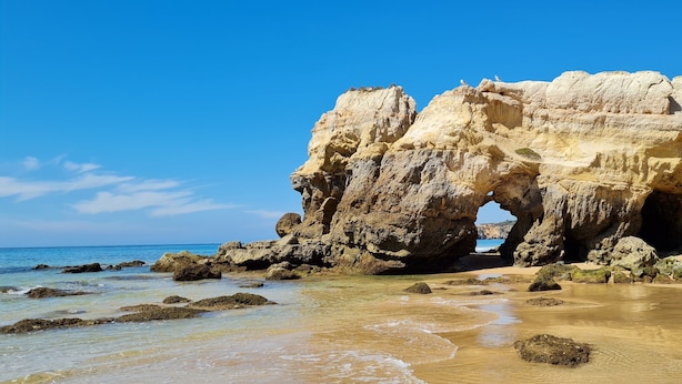 Formación rocosa costera con un arco natural, playa arenosa y cielo azul despejado.