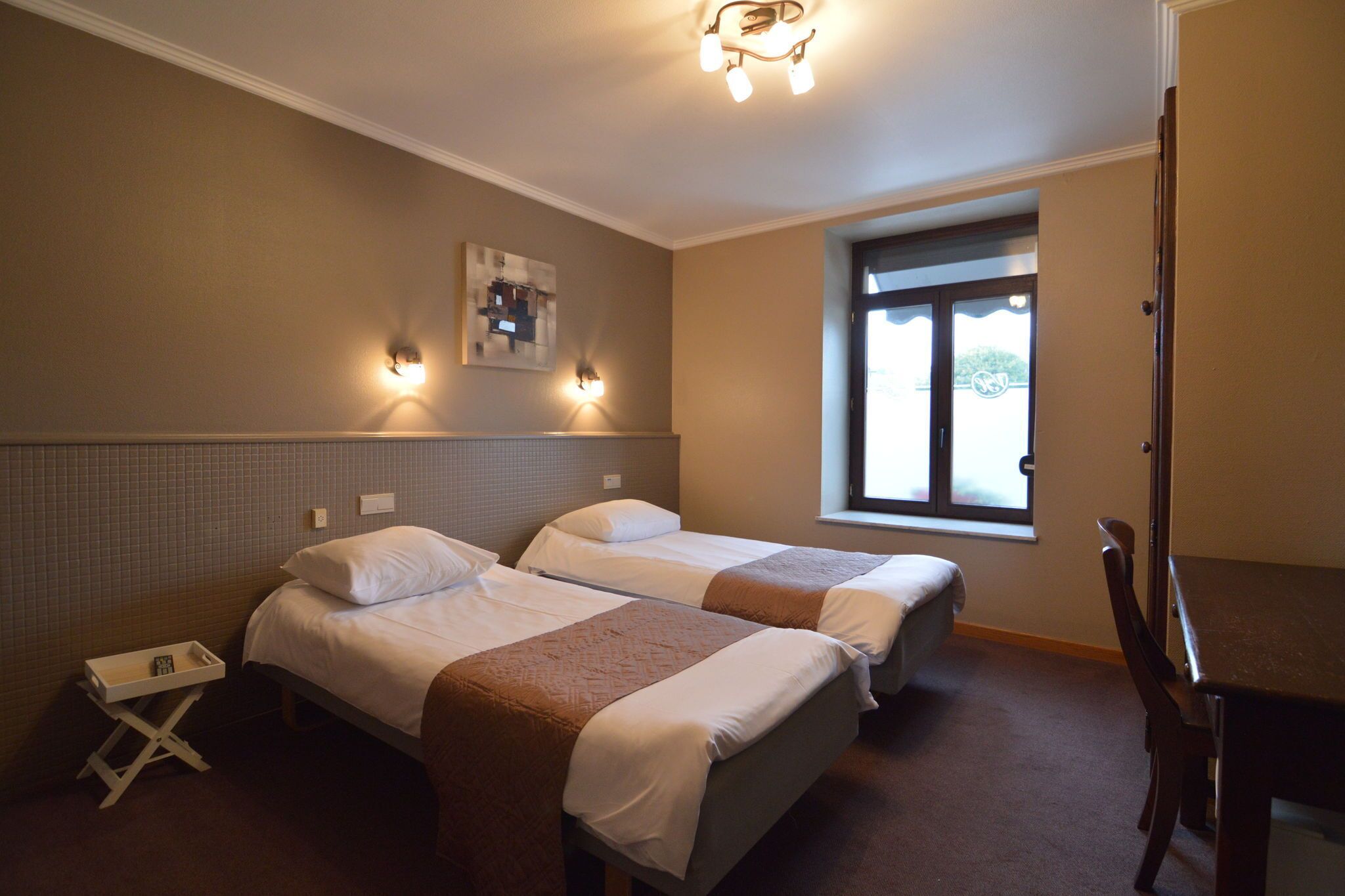 A hotel room with two beds, a small table, and a chair. There are wall sconces and a window with a view.