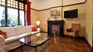 Una sala de estar con un sofá blanco, una mesa de centro de vidrio, una chimenea, un sillón de mimbre y un televisor montado en la pared.