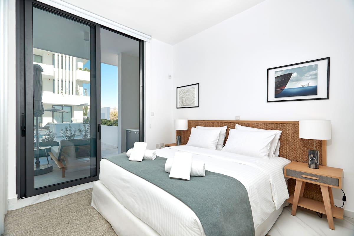 Habitación de hotel moderna con una cama grande, una mesita de noche con lámpara, un cuadro enmarcado en la pared y un ventanal corredero que da a un balcón.