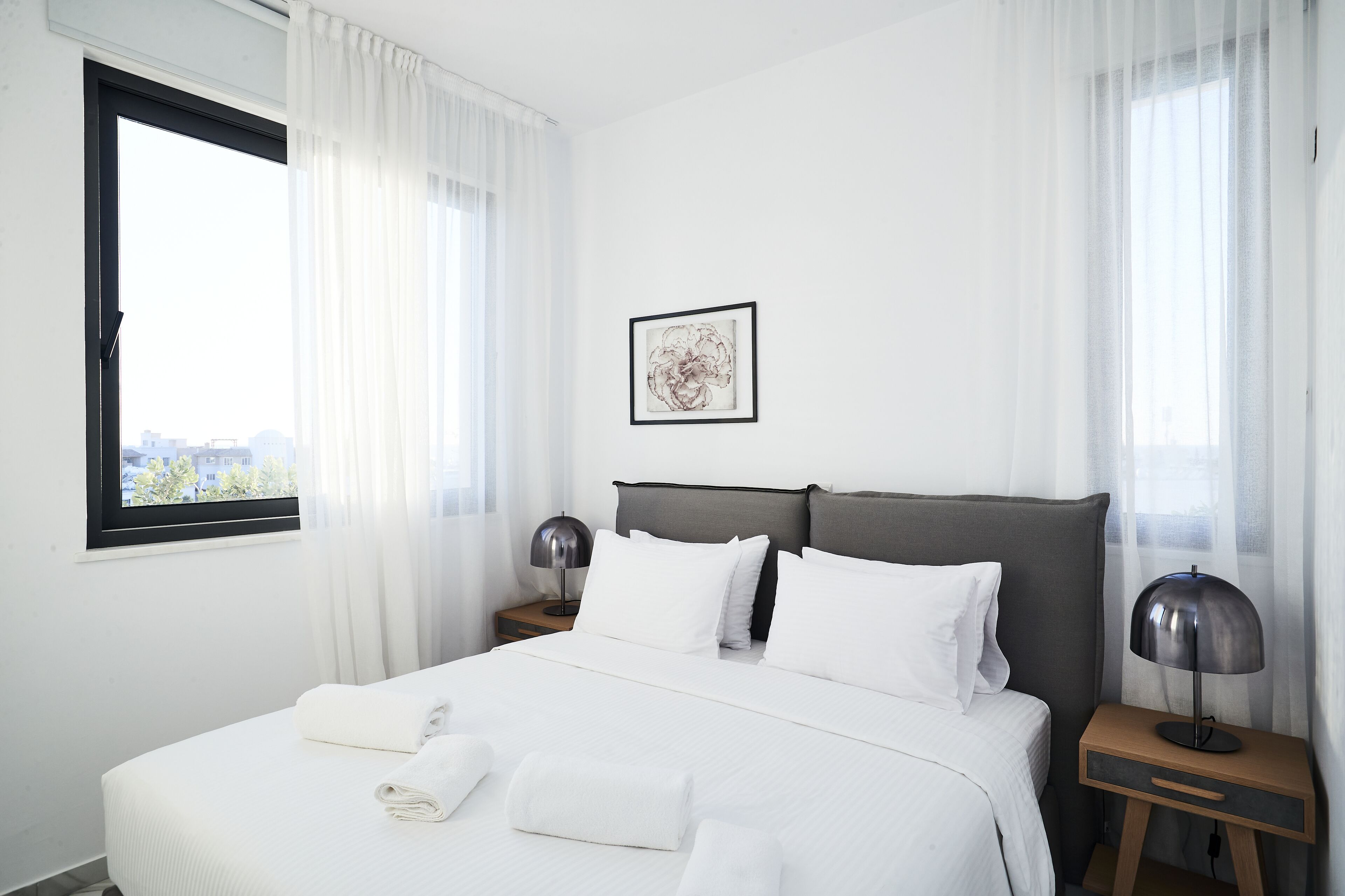 A hotel room with a large bed, white linens, a framed artwork on the wall, and a view of buildings through the window.