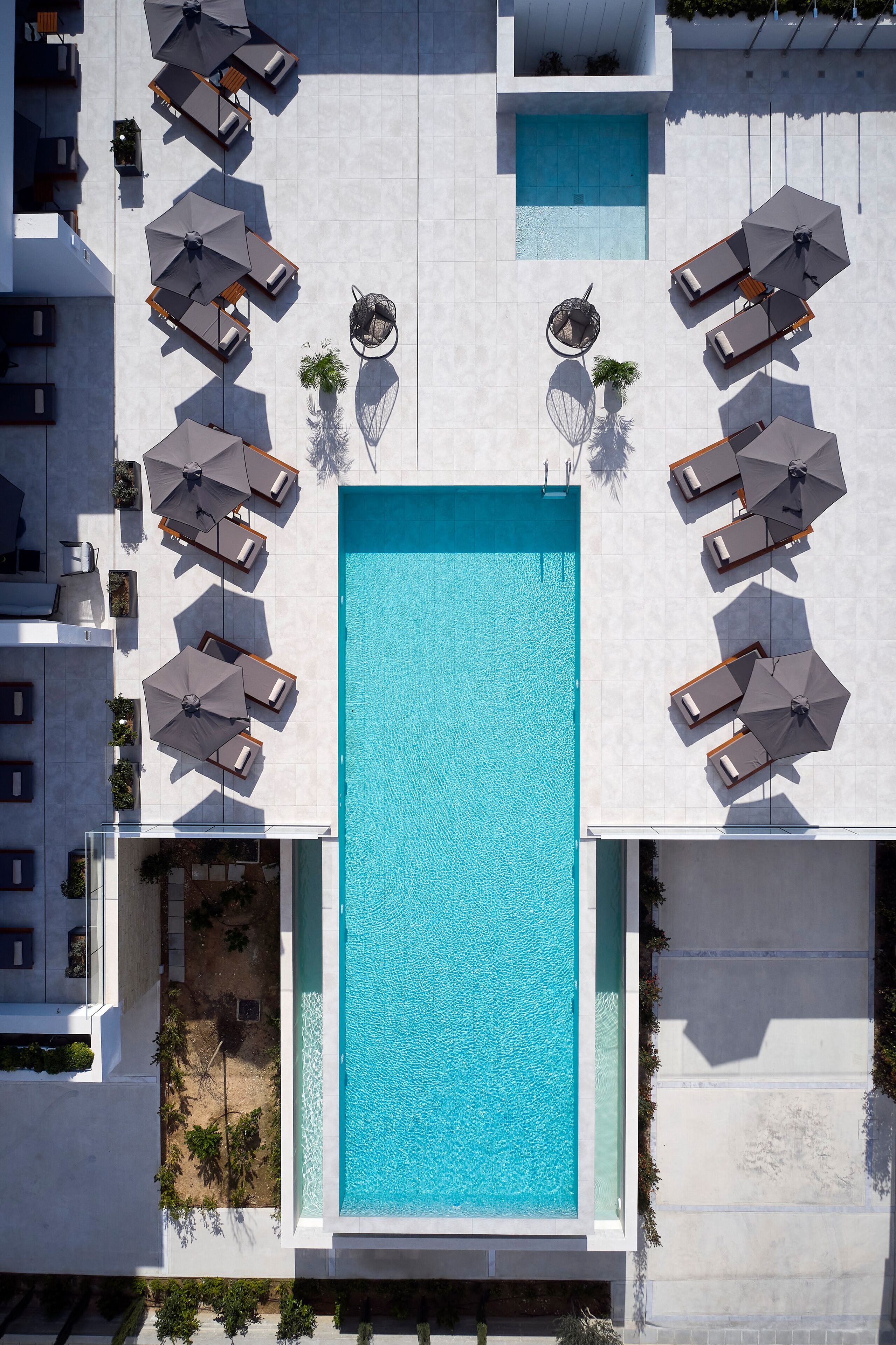 An aerial view of a modern hotel with a central swimming pool surrounded by lounge areas with umbrellas and seating.