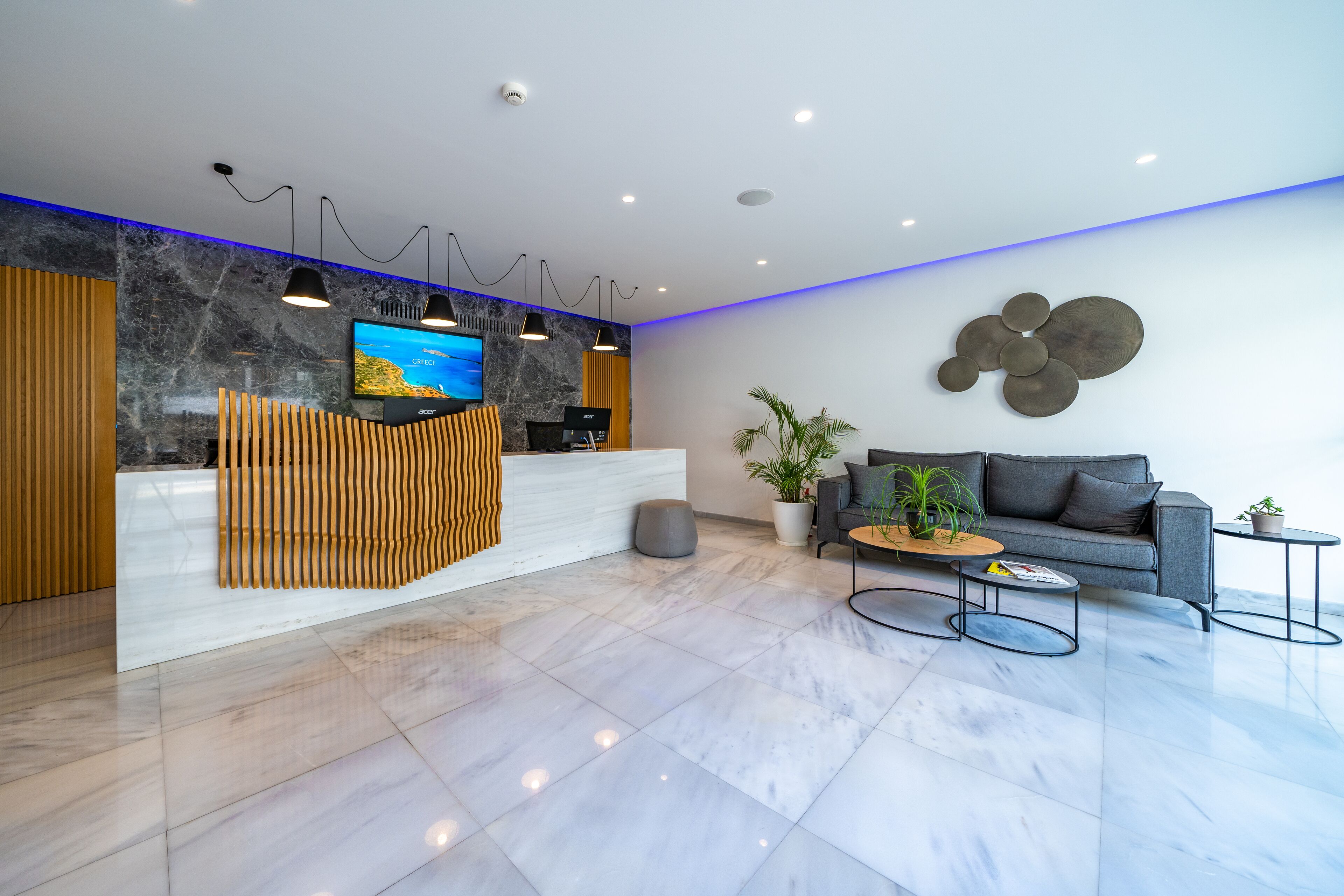 A modern reception area with a marble reception desk, a flat-screen TV, pendant lighting, a grey sofa, and a coffee table with magazines.