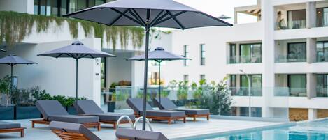 A poolside area with lounge chairs and umbrellas, a modern building with balconies, and a clear pool.