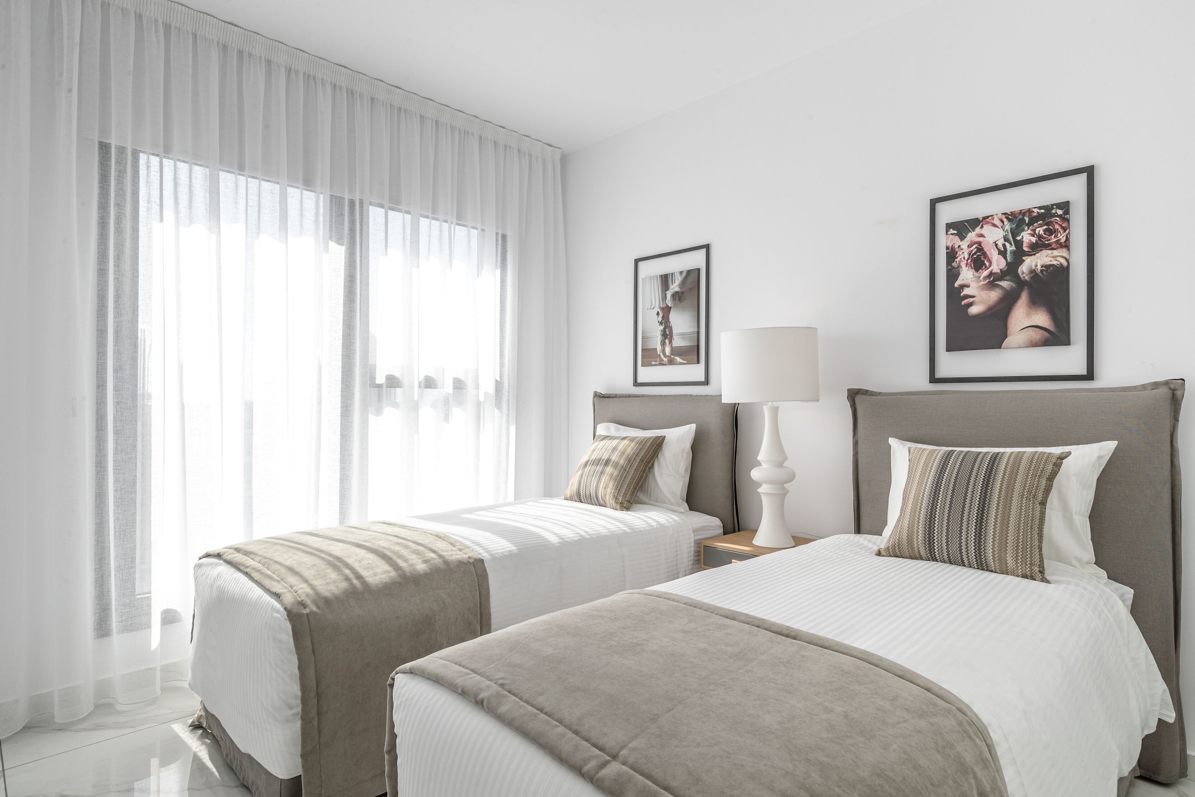 Una habitación de hotel con dos camas, un ventanal con cortinas translúcidas y dos cuadros en la pared.