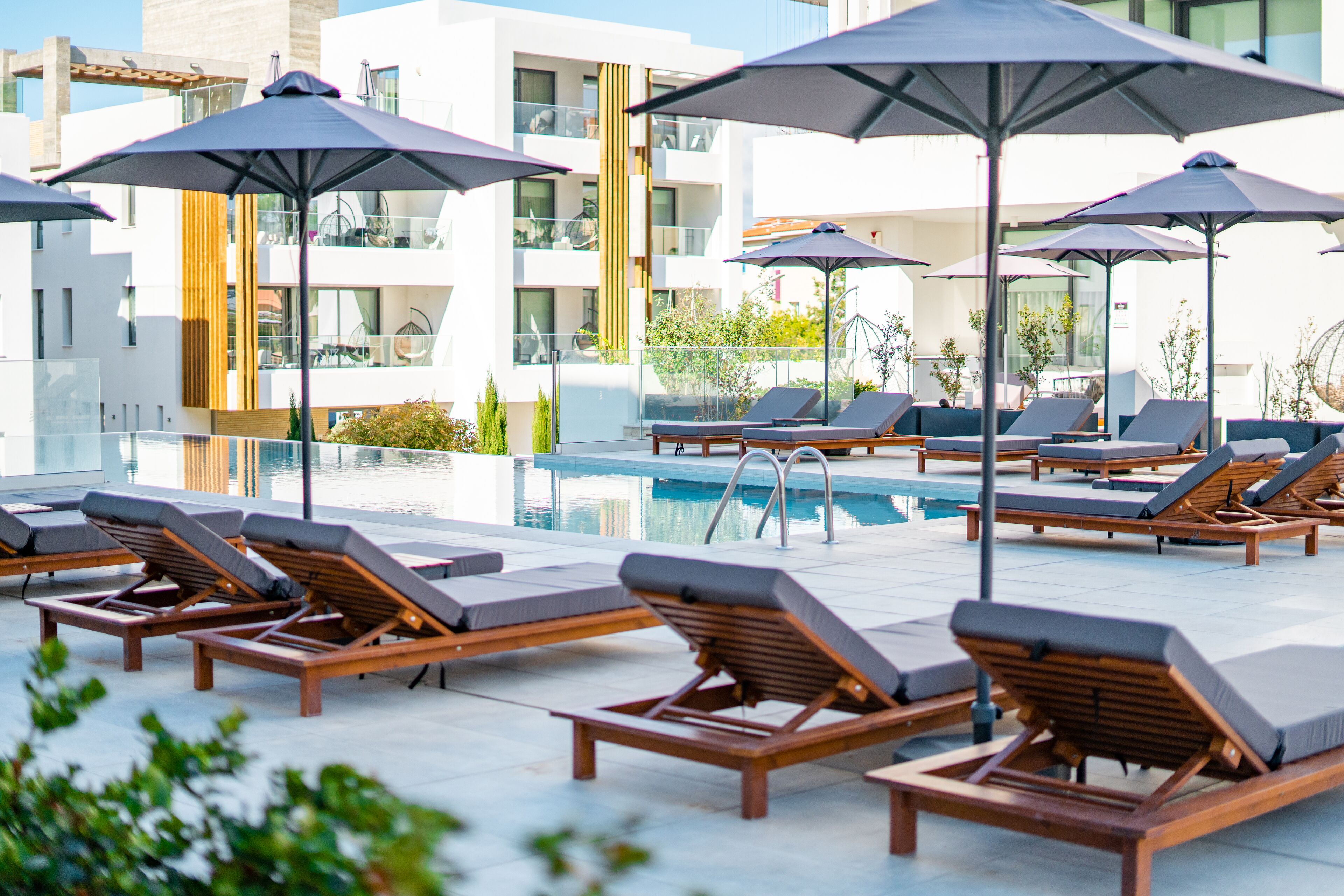 Área ao lado da piscina com espreguiçadeiras e guarda-sóis, piscina cristalina e edifícios modernos ao fundo.