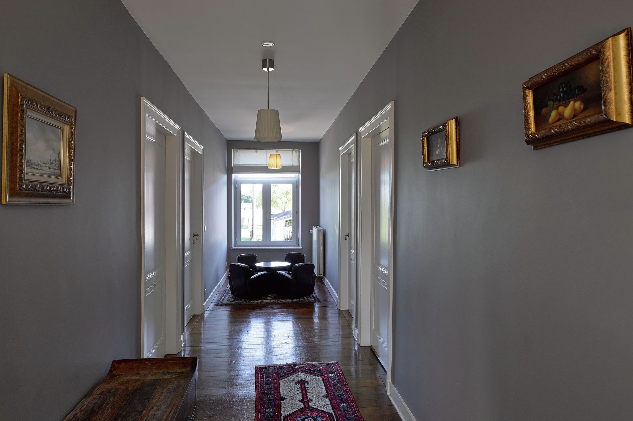 A hallway with a dining area at the end, featuring a chandelier, framed artwork, and a patterned rug.