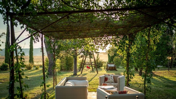 An outdoor seating area with wicker furniture under a pergola, overlooking a grassy field and a distant body of water.