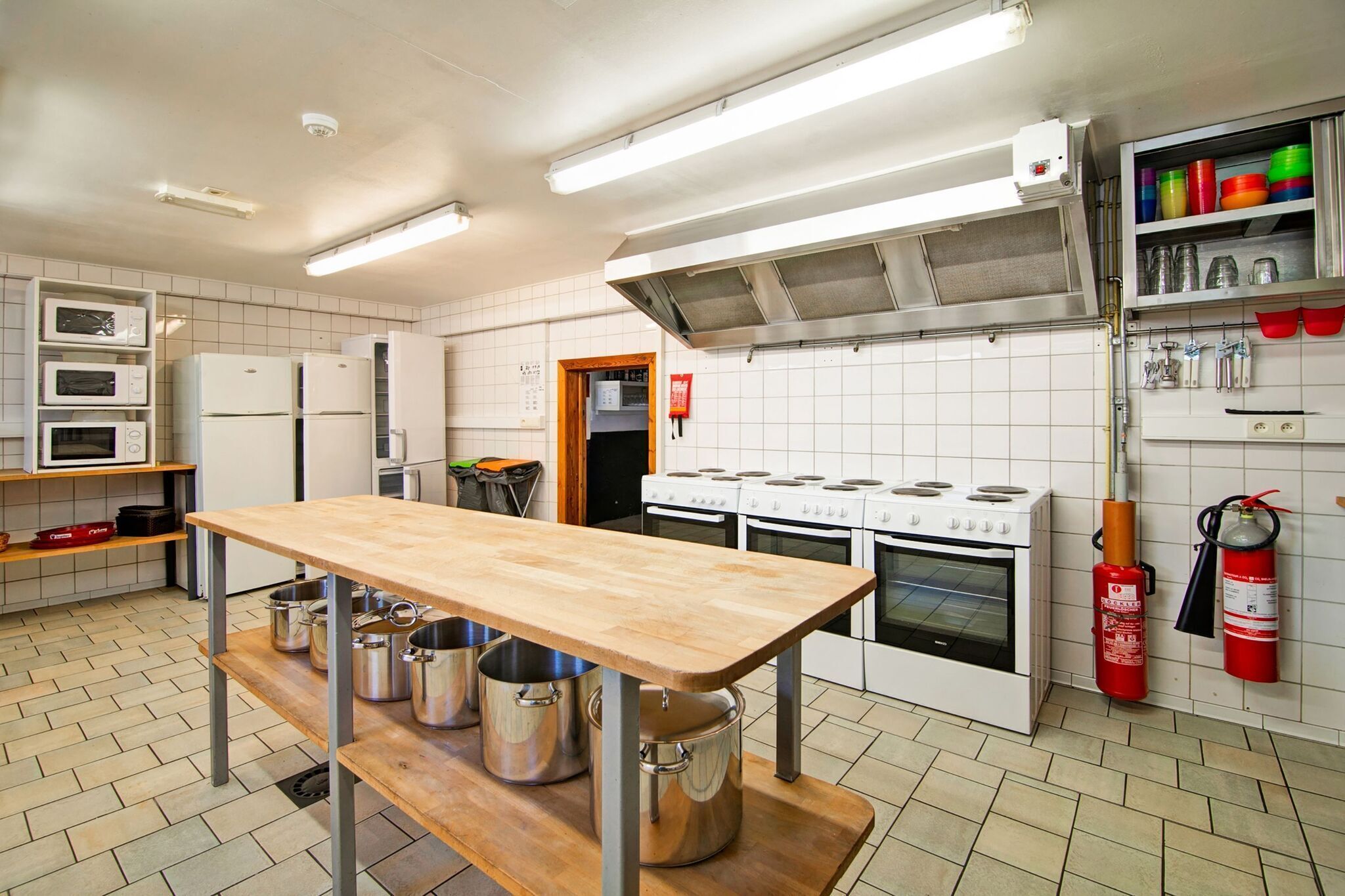 A kitchen with a wooden table, stainless steel pots, white tiled walls, a stove, and a fire extinguisher.