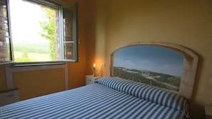 A bedroom with a bed, a nightstand, a lamp, a window with a view of greenery, and a mural on the wall. - Farmhouse in San Gimignano near Piazza Cisterna (San Gimignano)