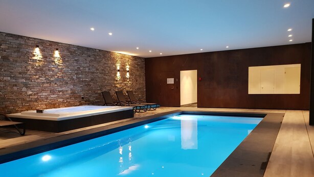 A modern indoor pool area with a stone wall, seating, and a hot tub. - Swanky Holiday Home in Waimes with Indoor Swimming Pool (Waimes)