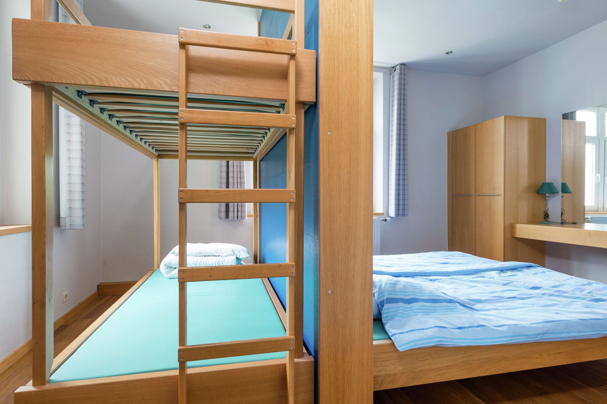 A bunk bed room with a wooden ladder, a single bed with a blue mattress, and a wooden wardrobe.