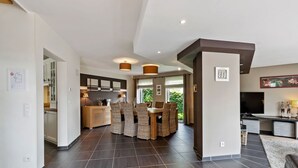 A modern kitchen with dark tiles, a dining area with wicker chairs, and a living space with a TV and artwork. - Holiday Home in Malmedy with Sauna & Bubble Bath (Malmedy)