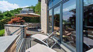 An outdoor patio area with modern furniture, a glass wall, and a view of the surrounding greenery. - Holiday Home in Malmedy with Sauna & Bubble Bath (Malmedy)