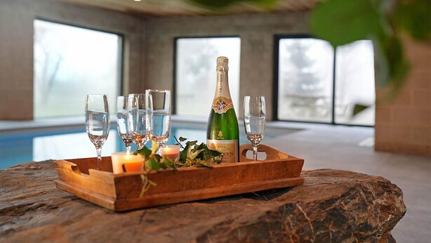 Een houten dienblad met champagne, glazen en een kaars op een stenen oppervlak in een kamer met grote ramen.