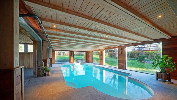 A large indoor swimming pool with a curved design, wooden ceiling beams, and large windows offering views of a garden.