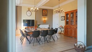 A dining area with a large table, chairs, and a chandelier. There is a television and a cabinet in the background.