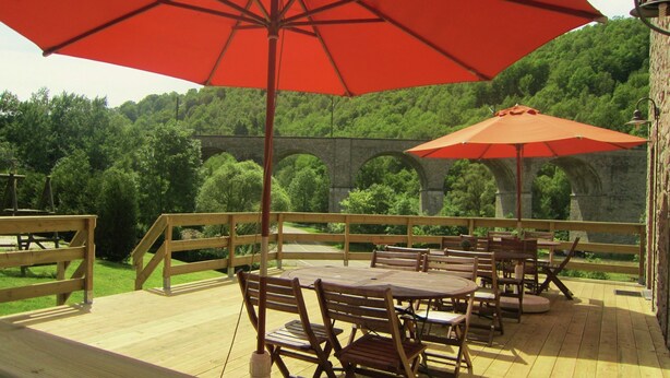 Eine Terrasse im Freien mit Holztischen und -stühlen, roten Sonnenschirmen und einer Steinbrücke im Hintergrund.