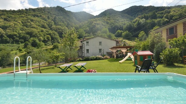 Une piscine dotée de chaises longues et d’un toboggan, entourée d’un paysage verdoyant luxuriant et avec une chaîne de montagnes en toile de fond.