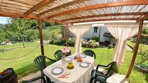 Un espace de restauration en plein air avec une table dressée pour quatre personnes, entourée de chaises vertes, sous une pergola en bois recouverte d’une nappe blanche.