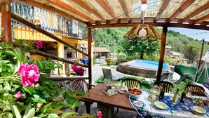Un espacio cubierto para comer al aire libre con una piscina al fondo, rodeado de follaje y un paisaje montañoso.