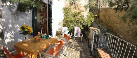 Un patio con una mesa lista para dos personas, rodeado de sillas, un pequeño jardín y un muro de piedra.