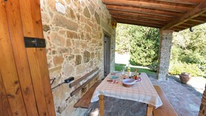 Une terrasse extérieure en pierre, avec une table et des bancs en bois, entourée de murs en pierre et d’une pergola en bois.