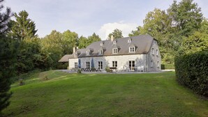 A stone building with multiple windows and a sloped roof, surrounded by a well-maintained lawn and trees.