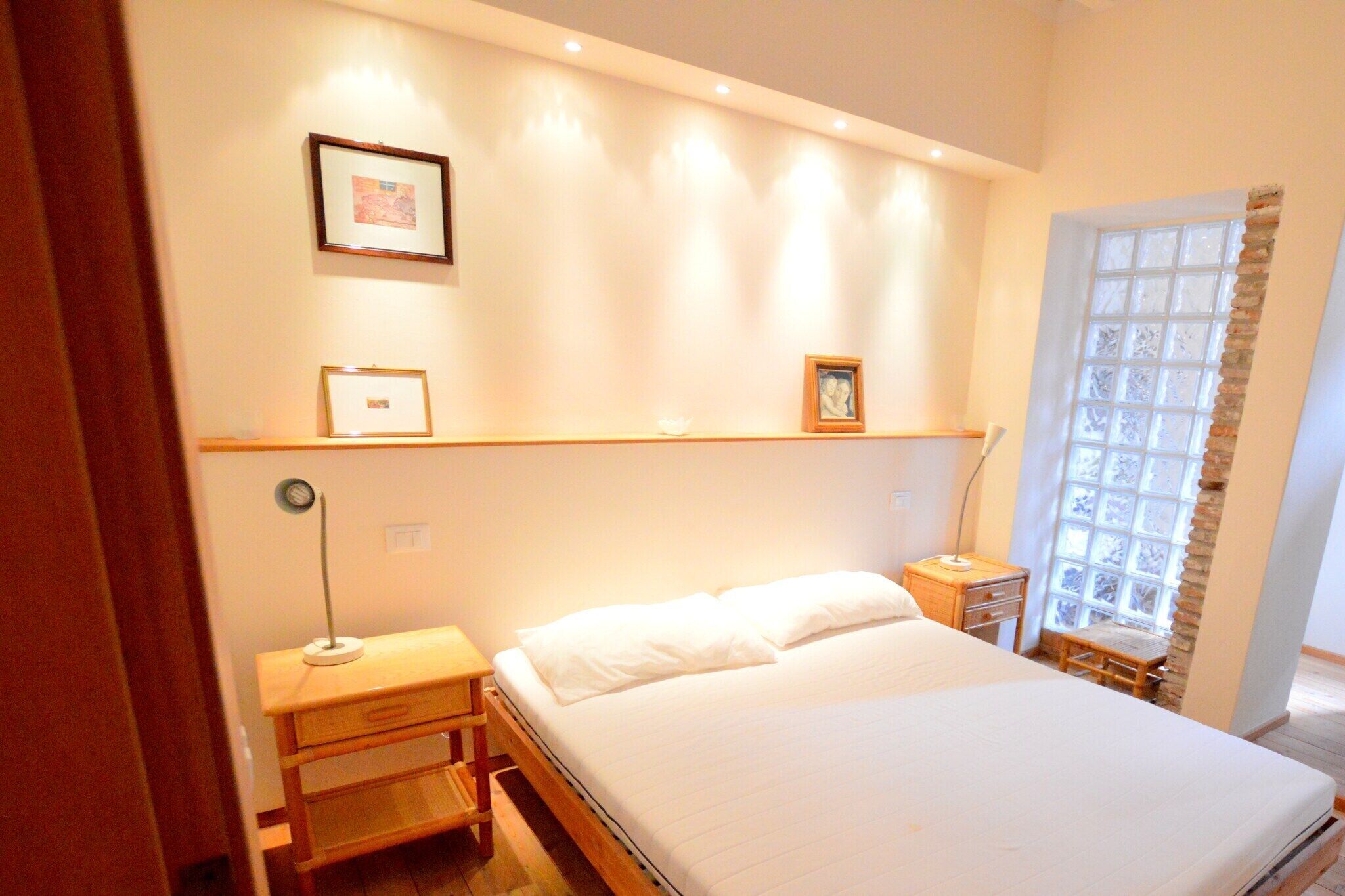 A bedroom with a bed, bedside tables, a nightstand, a window with a view, and wall-mounted shelves with framed pictures.
