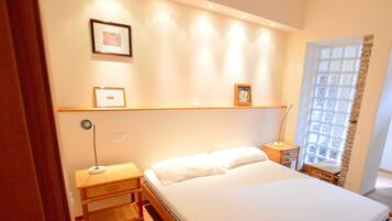 A bedroom with a bed, bedside tables, a nightstand, a window with a view, and wall-mounted shelves with framed pictures.