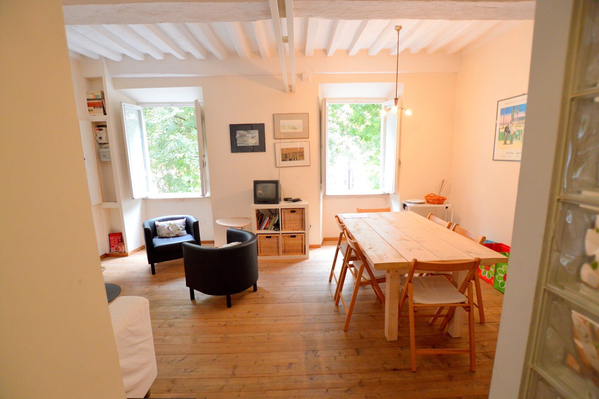 A room with a wooden floor, a dining table with chairs, a black armchair, a small white table, and a bookshelf.
