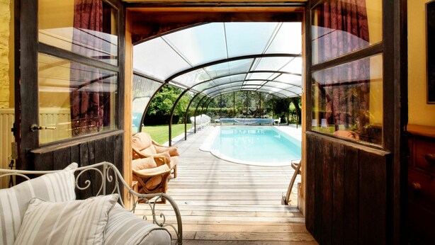 A covered outdoor area with a pool, wooden deck, and a seating area with chairs and a sofa. - Magnificent Holiday Home in Baronville with Heated Pool & Billiards (Beauraing Ardennes)