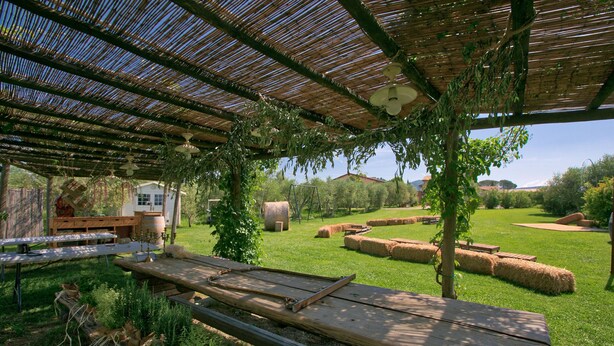 Un patio al aire libre con una pérgola, mesas de picnic, fardos de heno y un cobertizo pequeño.