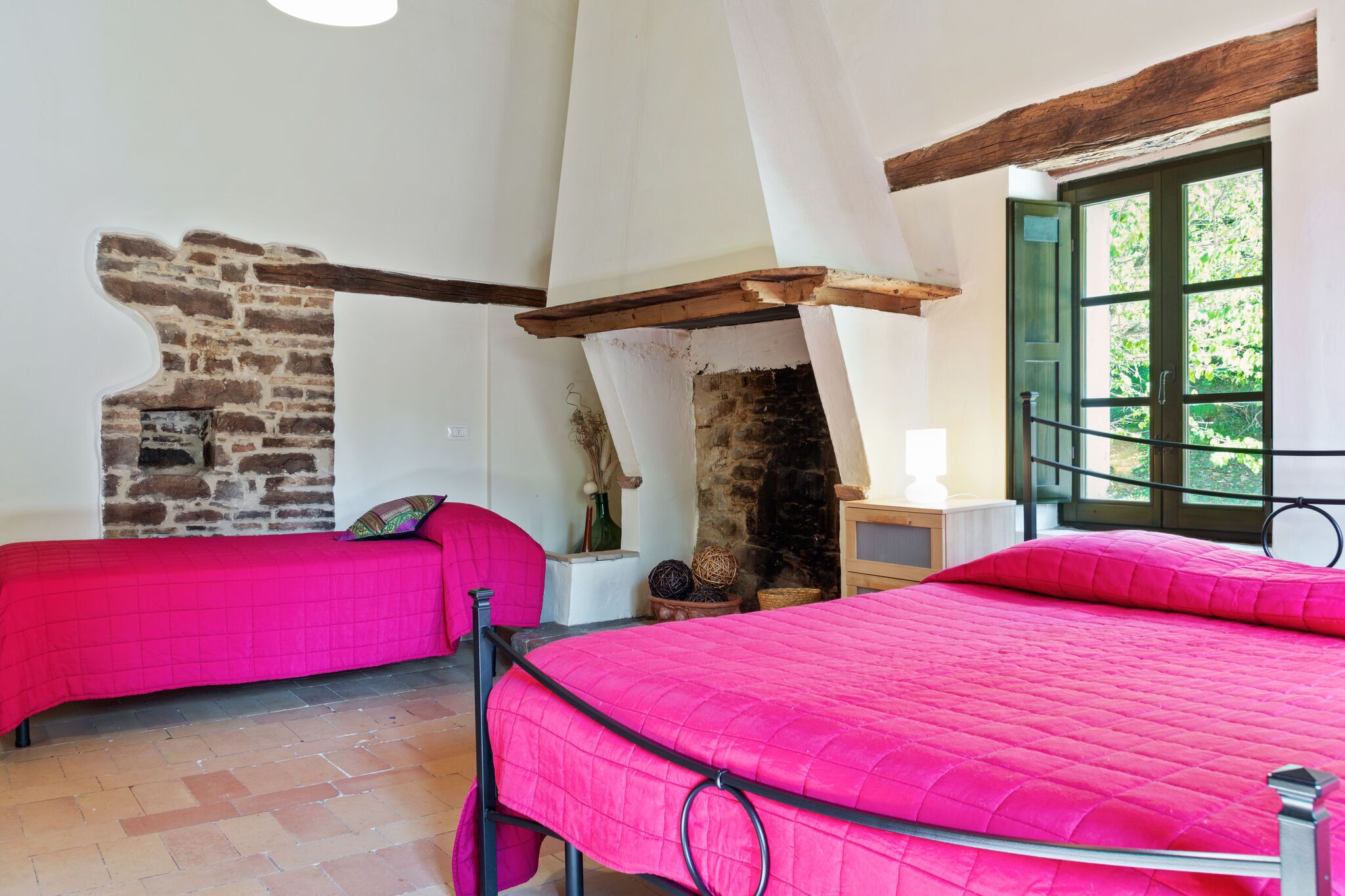 Una habitación con dos camas, pared de piedra, viga de madera y ventana con vista a follaje.