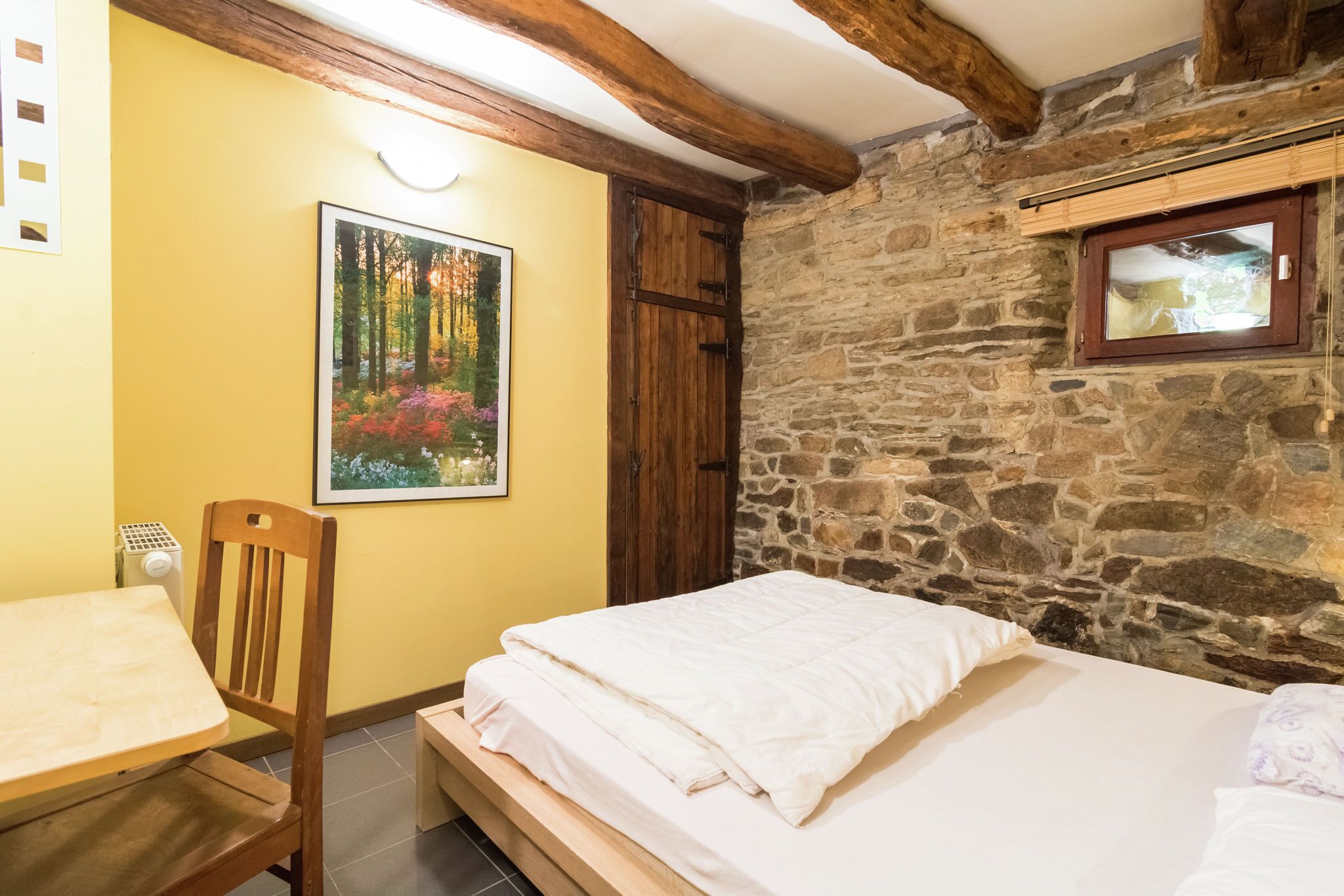 A bedroom with a bed, a wooden chair, a table, a painting, and a stone wall.