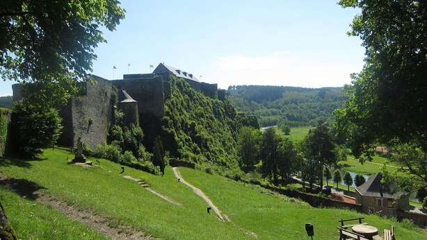 Um castelo numa colina com um caminho gramado a levá-lo, rodeado por árvores e um pequeno edifício com uma mesa de piquenique.