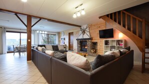 A spacious living room with a fireplace, a wooden staircase, and a dining area with a view. - Cottage in a well-equipped and comfortable farmhouse (Roy)
