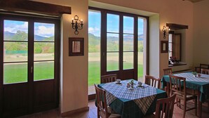 A dining area with large windows offering a view of a green landscape. There are tables set with tablecloths and chairs.