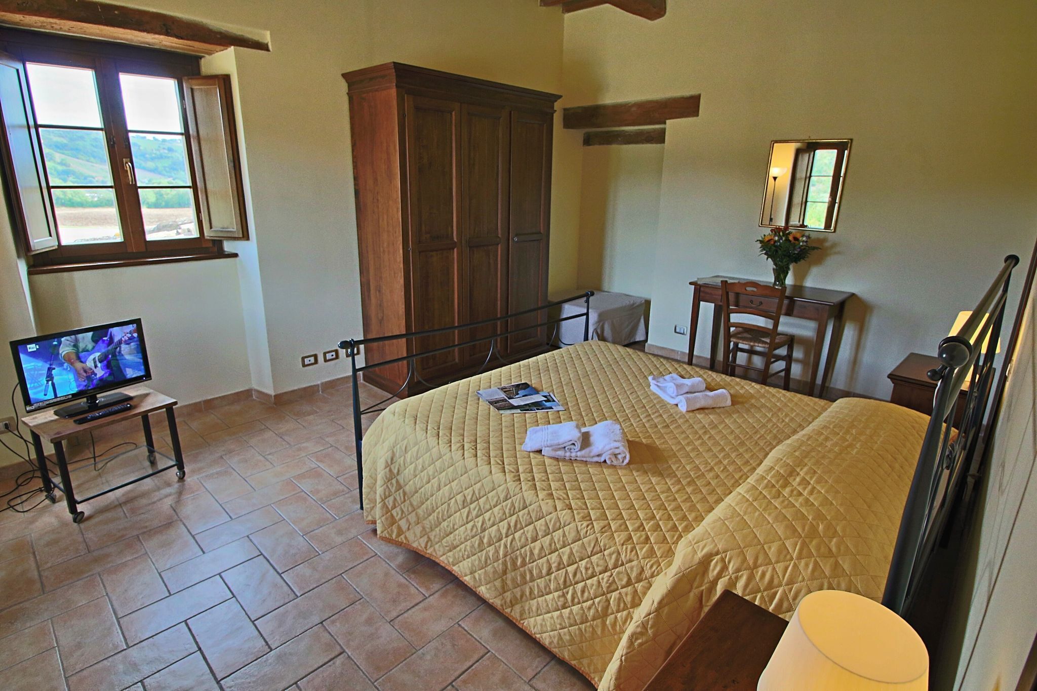 A bedroom with a bed, a television on a stand, a wooden wardrobe, a desk with a chair, and a window with a view of the outdoors.