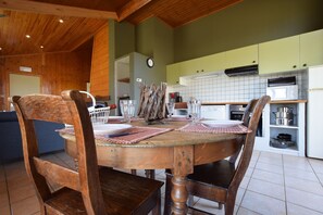 A wooden dining table with chairs, a kitchen area with cabinets and appliances, and a clock on the wall. - Holiday Home in Tenneville by Ourthe River (Tenneville)