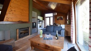 Een gezellige woonkamer met een open haard, een bank, een salontafel met glazen en een fles, en een keuken op de achtergrond.