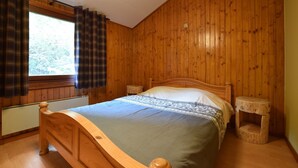 A wooden-paneled bedroom with a bed, a window with plaid curtains, and a small wooden stool.