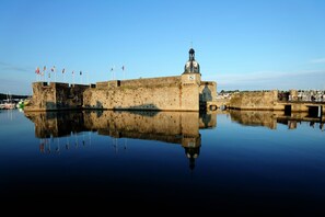 A historic stone fortress with a clock tower, flags, and a calm water body reflecting the structure.