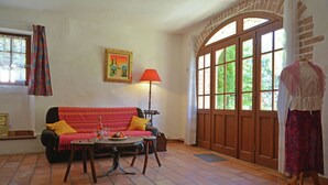 A cozy living room with a sofa, a table with a bottle and glasses, a lamp, and a large window with a view of greenery. - Villa in Bagard with Garden, BBQ, Garden Furniture, Playroom (Bagard)